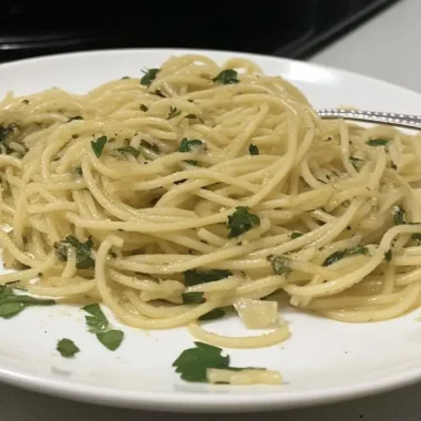 Plated spaghetti aglio e olio with golden olive oil coating pasta strands, fresh parsley garnish, and fork on white dinner plate