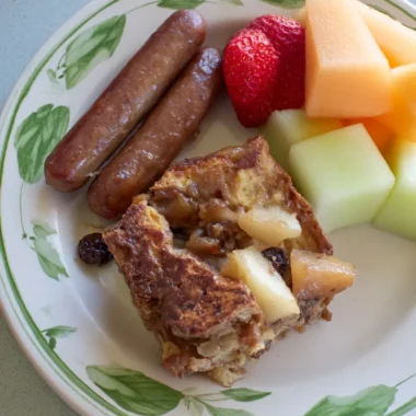 Warm slice of apple-raisin French toast casserole served with breakfast sausages, strawberries, cantaloupe, and honeydew melon on a white plate with green leaf trim.