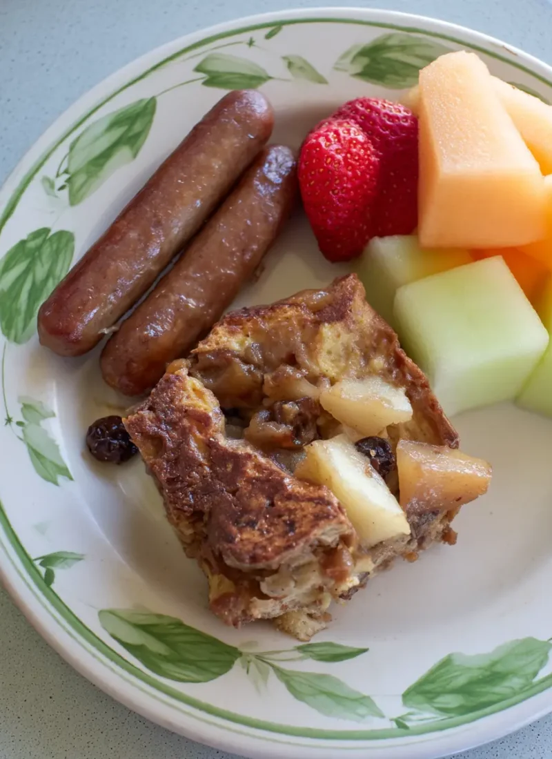 Warm slice of apple-raisin French toast casserole served with breakfast sausages, strawberries, cantaloupe, and honeydew melon on a white plate with green leaf trim.