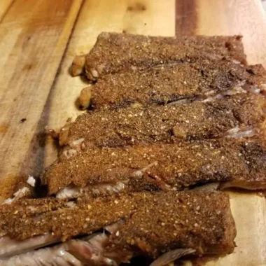 Sliced baked baby back ribs with dry rub resting on a wooden cutting board.