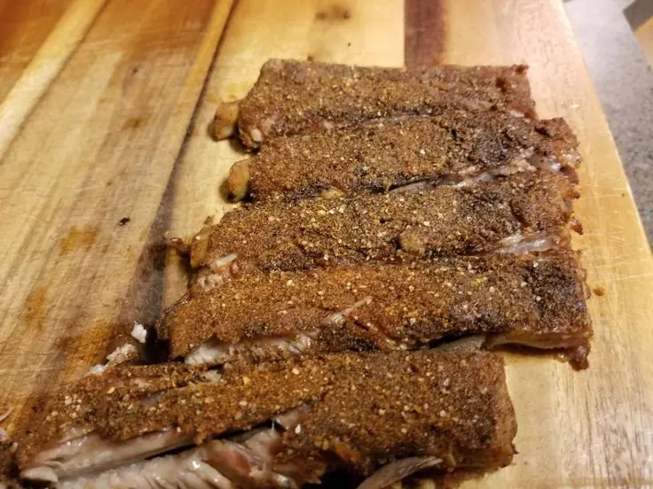 Sliced baked baby back ribs with dry rub resting on a wooden cutting board.