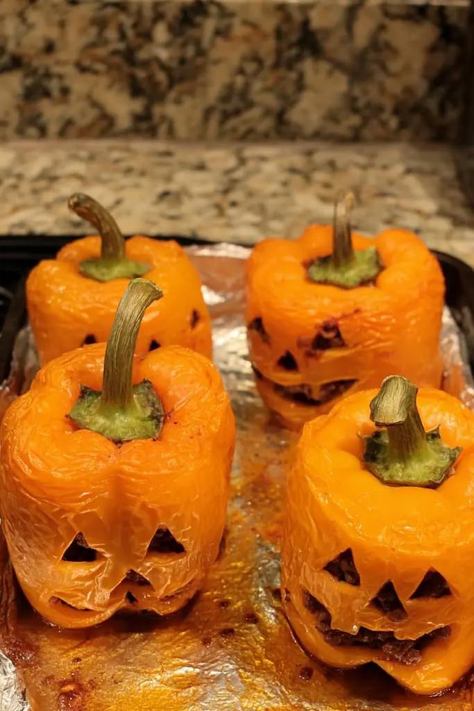 Four roasted orange bell peppers with carved jack-o'-lantern faces on a foil-lined baking sheet, their skins softened and slightly wrinkled from baking, with caramelized juices pooling beneath