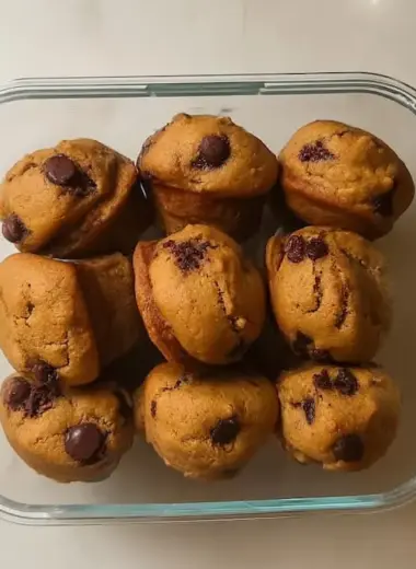 Nine golden banana protein muffins with chocolate chips arranged in clear glass storage container viewed from above