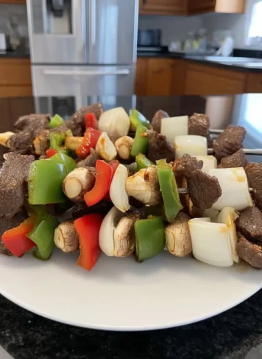 Uncooked beef kabobs threaded with steak cubes, red and green bell peppers, onion chunks, and button mushrooms on a white plate in a home kitchen.