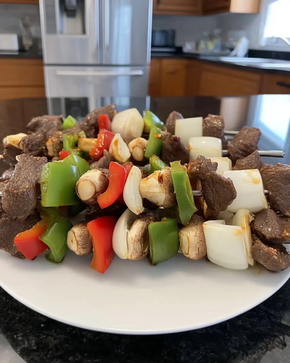 Uncooked beef kabobs threaded with steak cubes, red and green bell peppers, onion chunks, and button mushrooms on a white plate in a home kitchen.