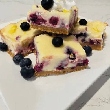Creamy blueberry cream cheese bars with a golden graham cracker crust, topped with fresh blueberries, whipped cream, and lemon zest on a white plate.
