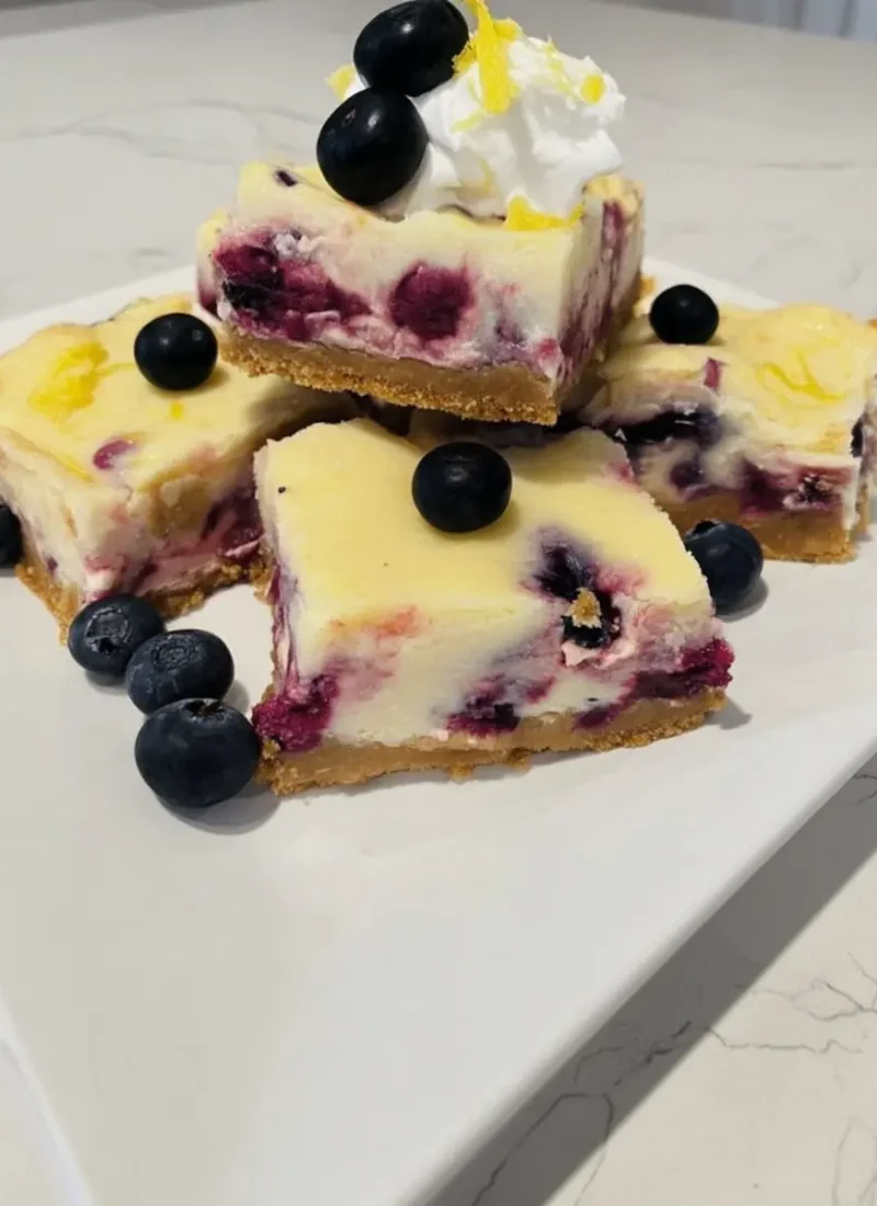 Creamy blueberry cream cheese bars with a golden graham cracker crust, topped with fresh blueberries, whipped cream, and lemon zest on a white plate.