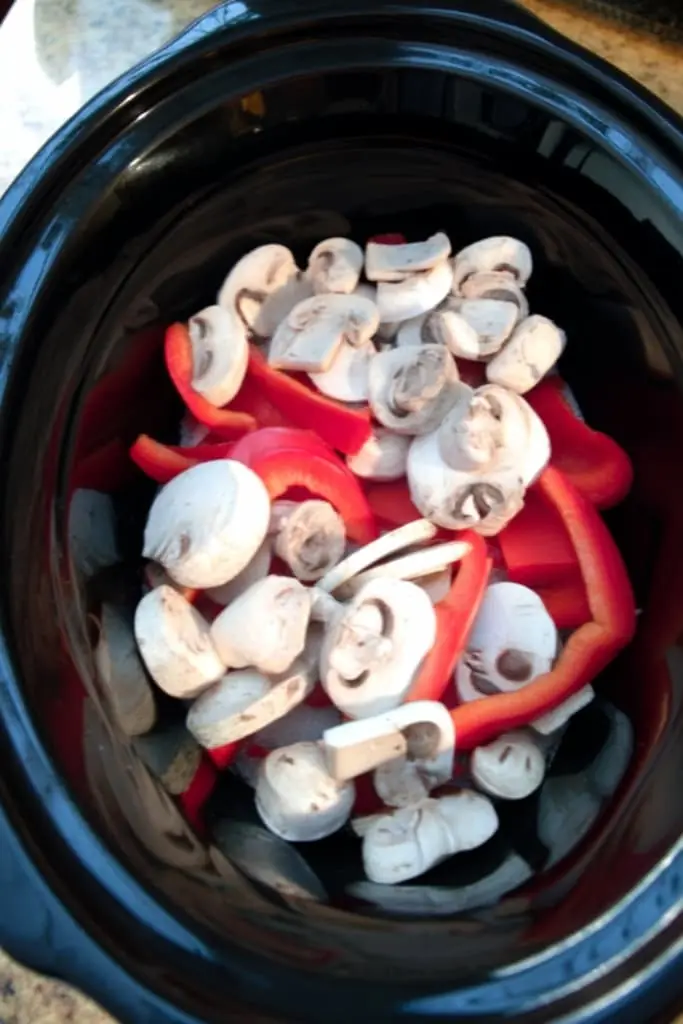 Fresh sliced mushrooms and red bell pepper strips layered in black slow cooker insert for chicken cacciatore recipe preparation