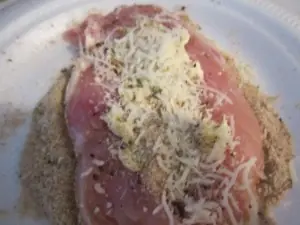 Uncooked skinless chicken breast laid flat, topped with shredded mozzarella cheese, breadcrumbs, herbs, and spices on a white plate