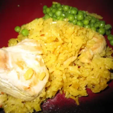 Close-up of homemade Arroz Con Pollo with yellow rice, tender chicken pieces, and a side of green peas on a red plate