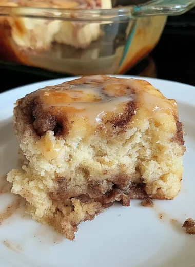 Close-up of a soft cinnamon roll cake slice with gooey cinnamon swirls and vanilla glaze, served on a white plate.