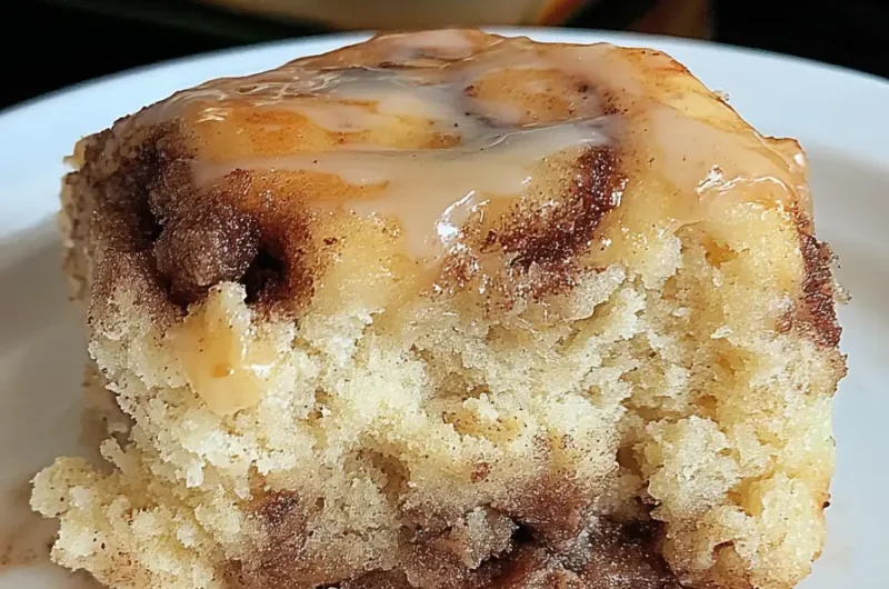Close-up of a soft cinnamon roll cake slice with gooey cinnamon swirls and vanilla glaze, served on a white plate.