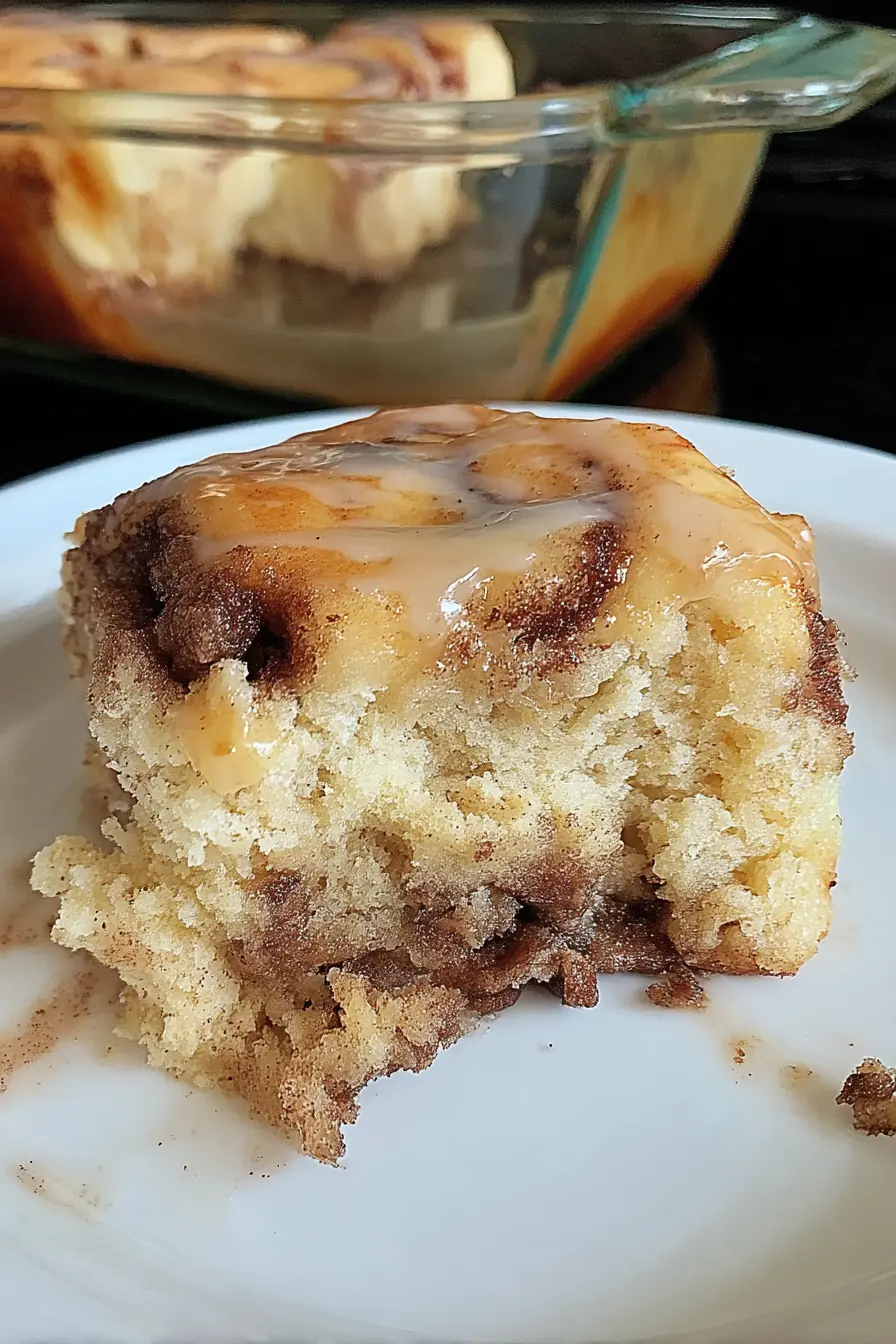 Close-up of a soft cinnamon roll cake slice with gooey cinnamon swirls and vanilla glaze, served on a white plate.