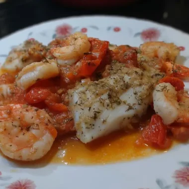 Close-up of baked cod and shrimp in golden garlic tomato sauce on a floral plate, seasoned with dried oregano and black pepper