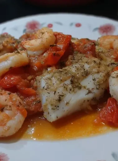 Close-up of baked cod and shrimp in golden garlic tomato sauce on a floral plate, seasoned with dried oregano and black pepper
