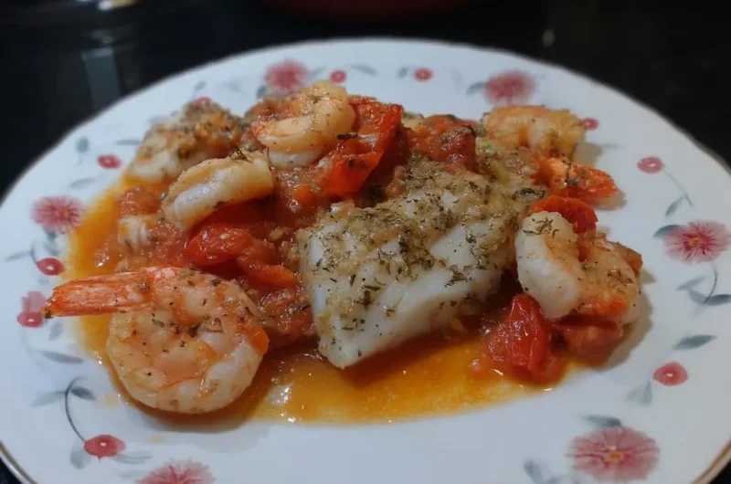 Close-up of baked cod and shrimp in golden garlic tomato sauce on a floral plate, seasoned with dried oregano and black pepper