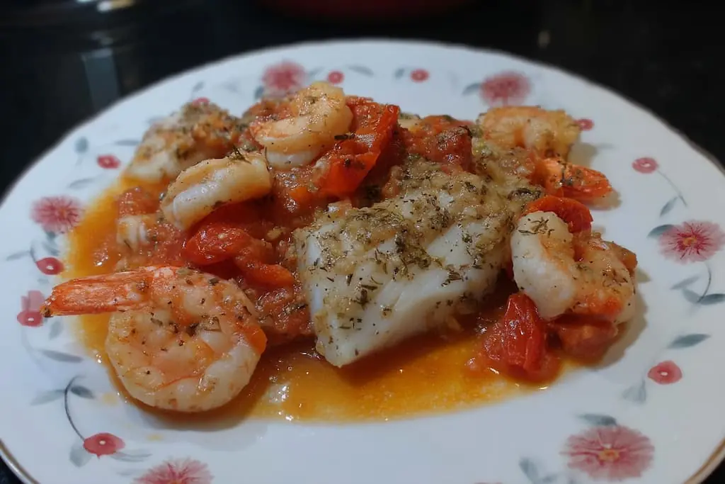 Close-up of baked cod and shrimp in golden garlic tomato sauce on a floral plate, seasoned with dried oregano and black pepper