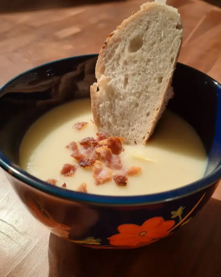 Creamy potato leek soup in dark blue bowl topped with crispy bacon bits and served with crusty artisan bread slice