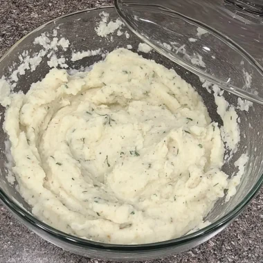 Bowl of creamy roasted garlic mashed potatoes dotted with fresh herbs on a speckled kitchen countertop