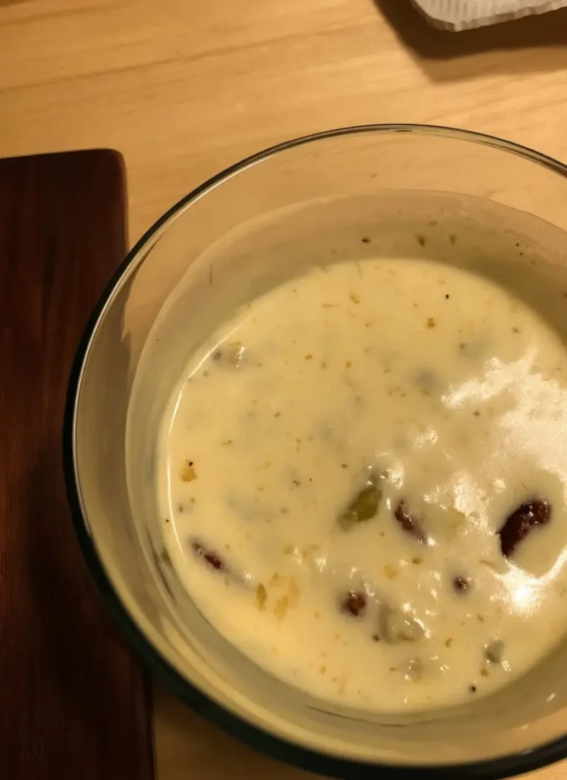 Close-up of creamy white queso dip with mild peppers and beans in a glass bowl on a wooden table.