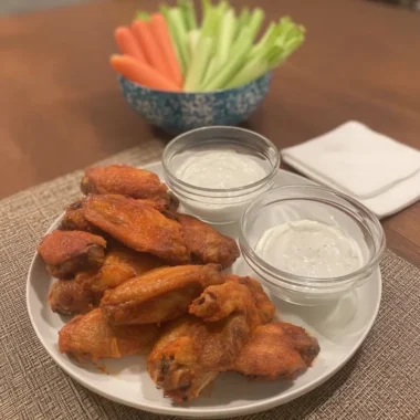 Close-up of crispy oven-baked Buffalo wings coated in a spicy sauce, arranged on a white plate with two small bowls of creamy ranch dressing; a blurred bowl of celery and carrot sticks sits in the background on a wooden table.