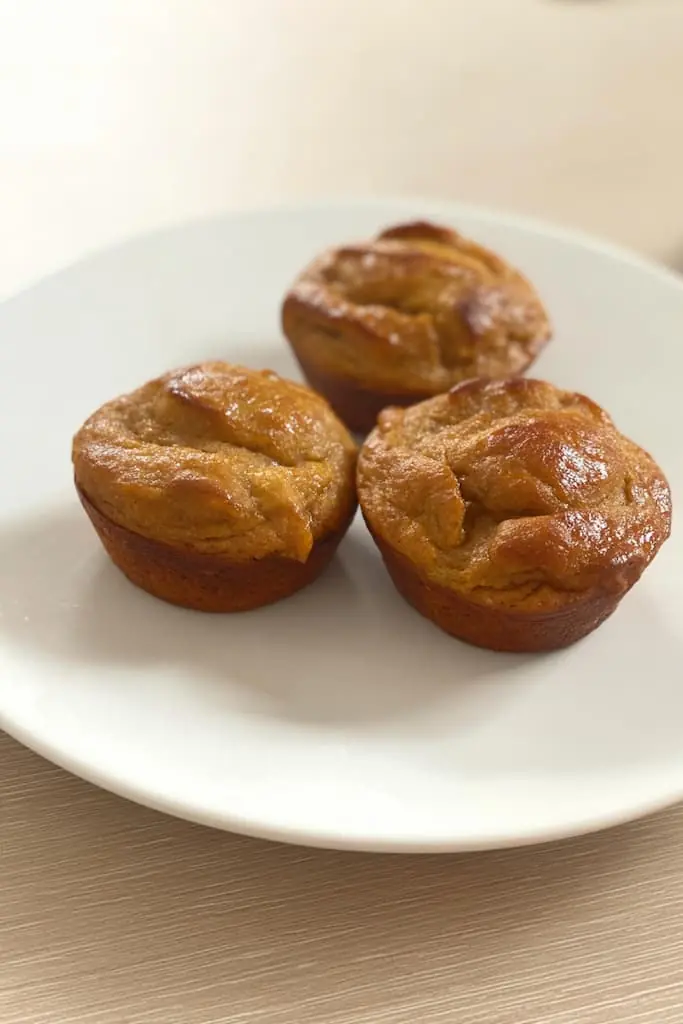 Golden brown high protein banana muffins on a white plate showing their perfectly risen tops and glossy finish from maple syrup drizzle