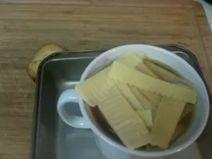 White crock of French onion soup topped with toasted baguette and raw Gruyere strips before broiling