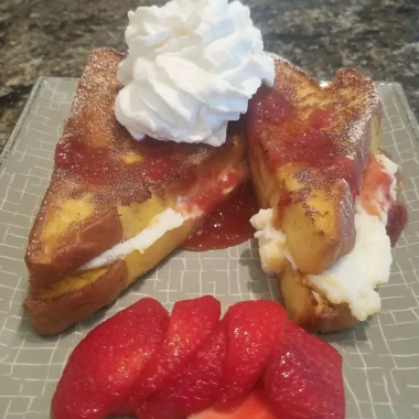 Fried strawberry cheesecake sandwiches with whipped cream on top, strawberry sauce, and fresh sliced strawberries on the side, served on a gray patterned plate.