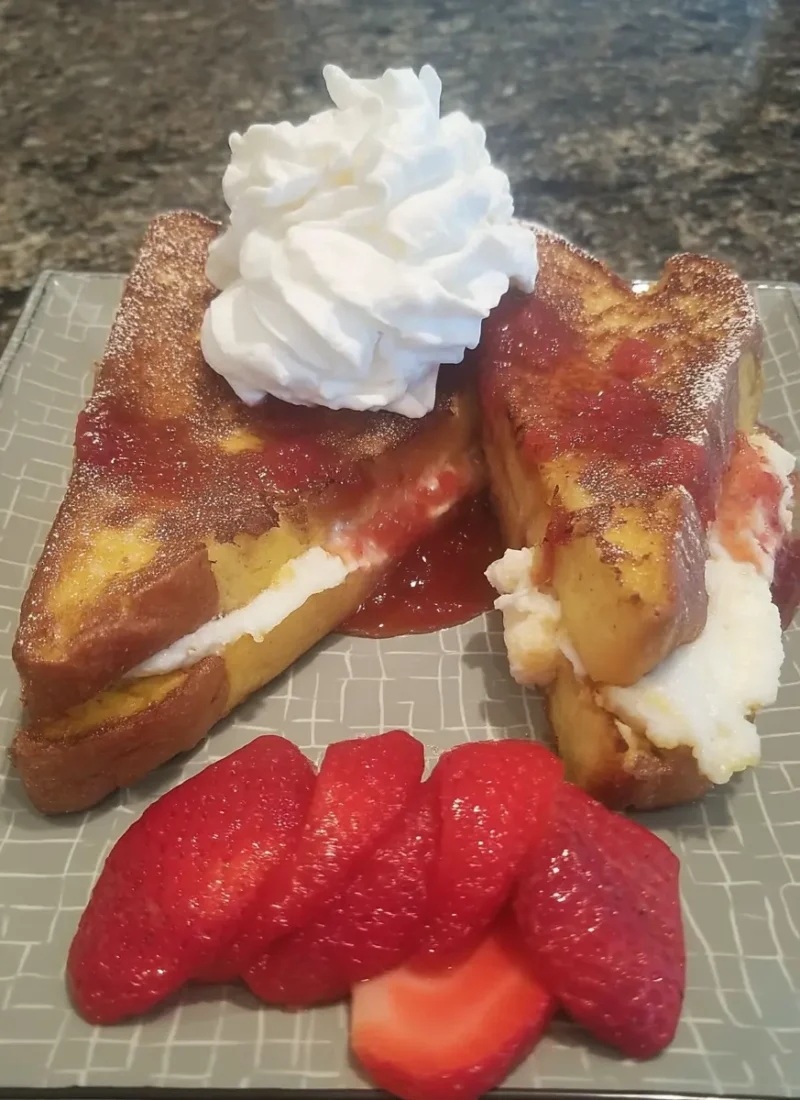 Fried strawberry cheesecake sandwiches with whipped cream on top, strawberry sauce, and fresh sliced strawberries on the side, served on a gray patterned plate.