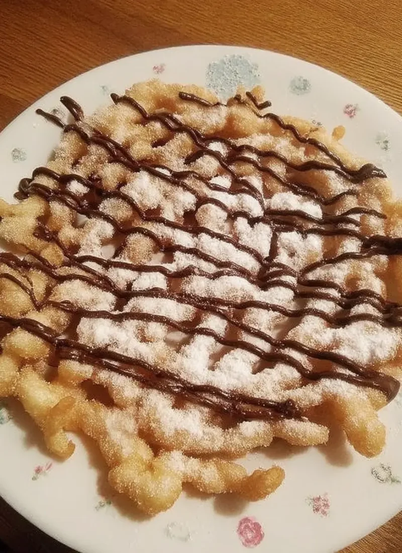 Golden brown funnel cake bites topped with powdered sugar and a generous drizzle of chocolate syrup, served on a floral-patterned plate.