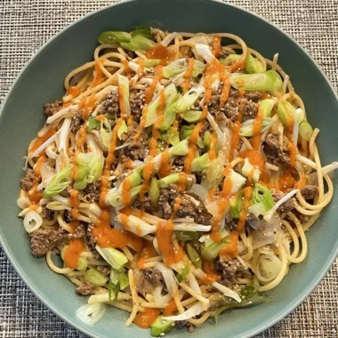 Bowl of Asian-style garlic noodles with seasoned ground beef, bean sprouts, sliced green onions, sesame seeds, and a drizzle of spicy orange chili sauce.