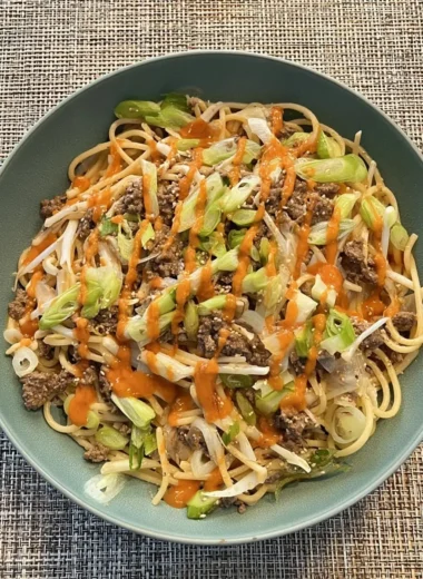 Bowl of Asian-style garlic noodles with seasoned ground beef, bean sprouts, sliced green onions, sesame seeds, and a drizzle of spicy orange chili sauce.