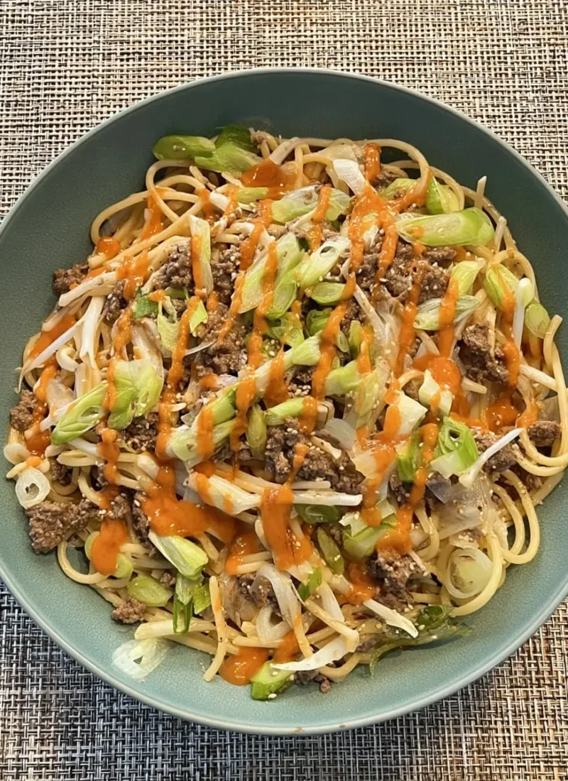 Bowl of Asian-style garlic noodles with seasoned ground beef, bean sprouts, sliced green onions, sesame seeds, and a drizzle of spicy orange chili sauce.