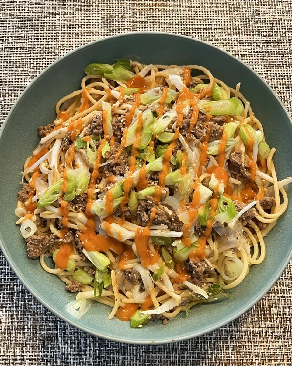 Bowl of Asian-style garlic noodles with seasoned ground beef, bean sprouts, sliced green onions, sesame seeds, and a drizzle of spicy orange chili sauce.