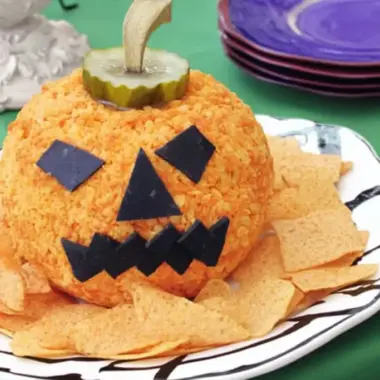 Jack-o'-lantern cheese ball with black olive square eyes and jagged mouth on white decorative plate with tortilla chips and purple plates in background