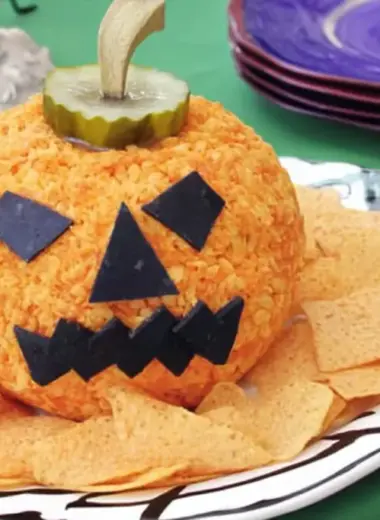Jack-o'-lantern cheese ball with black olive square eyes and jagged mouth on white decorative plate with tortilla chips and purple plates in background