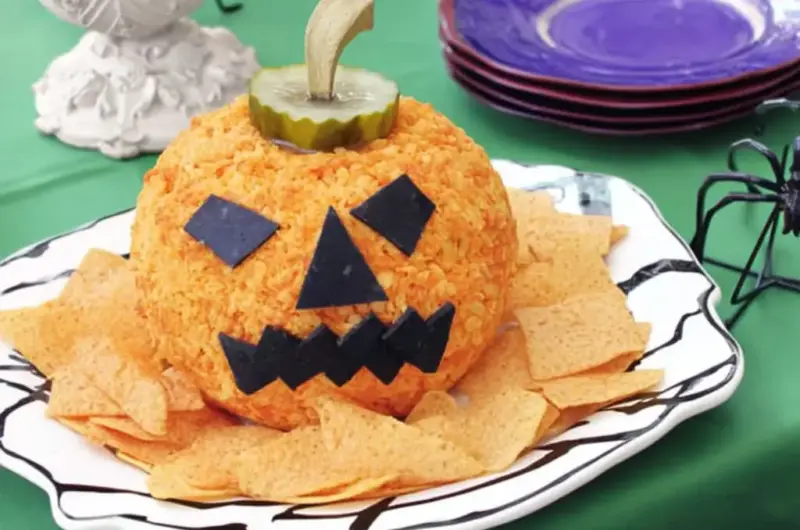 Jack-o'-lantern cheese ball with black olive square eyes and jagged mouth on white decorative plate with tortilla chips and purple plates in background