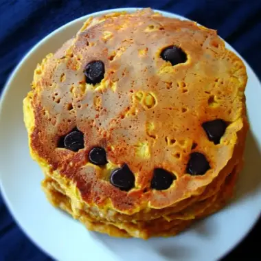 Golden pumpkin pancake on white plate with chocolate chip jack-o-lantern face showing triangle eyes and wavy smile
