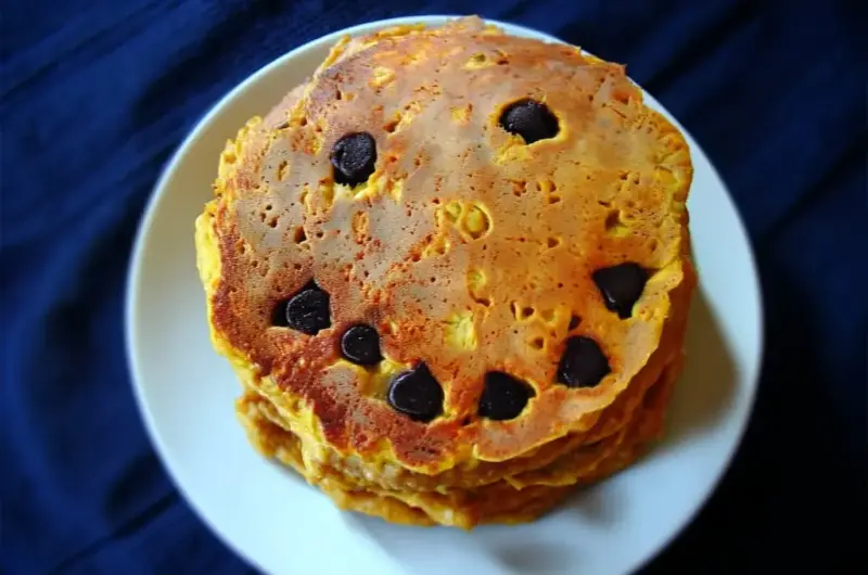 Golden pumpkin pancake on white plate with chocolate chip jack-o-lantern face showing triangle eyes and wavy smile