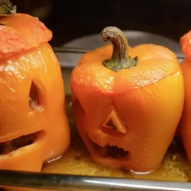 Tender roasted jack-o'-lantern bell peppers with carved faces in a metal baking dish, juices pooling around the bases after one hour of baking