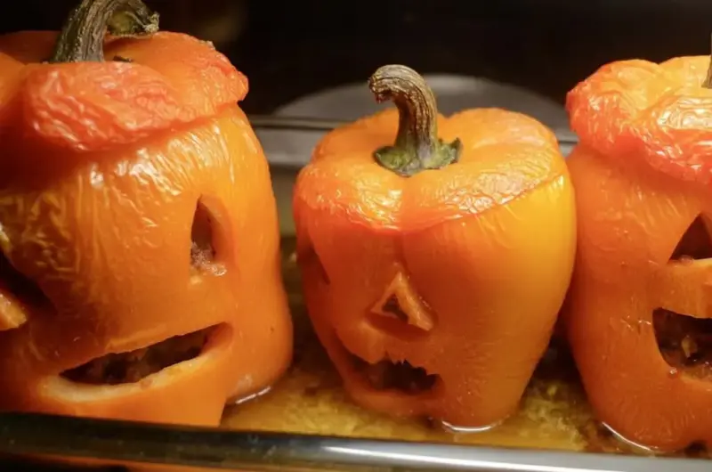 Tender roasted jack-o'-lantern bell peppers with carved faces in a metal baking dish, juices pooling around the bases after one hour of baking