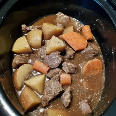 Slow cooker beef stew in a black crockpot, showing tender beef chunks, potato wedges, and carrot pieces in a thick brown gravy.