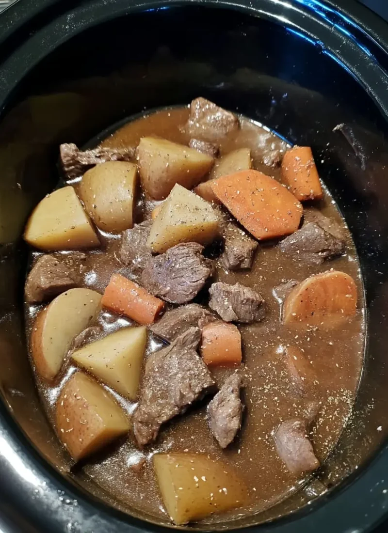 Slow cooker beef stew in a black crockpot, showing tender beef chunks, potato wedges, and carrot pieces in a thick brown gravy.