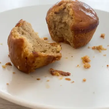 Cross-section view of a high protein banana muffin showing the tender, golden crumb structure with one half broken apart on a white plate with scattered crumbs