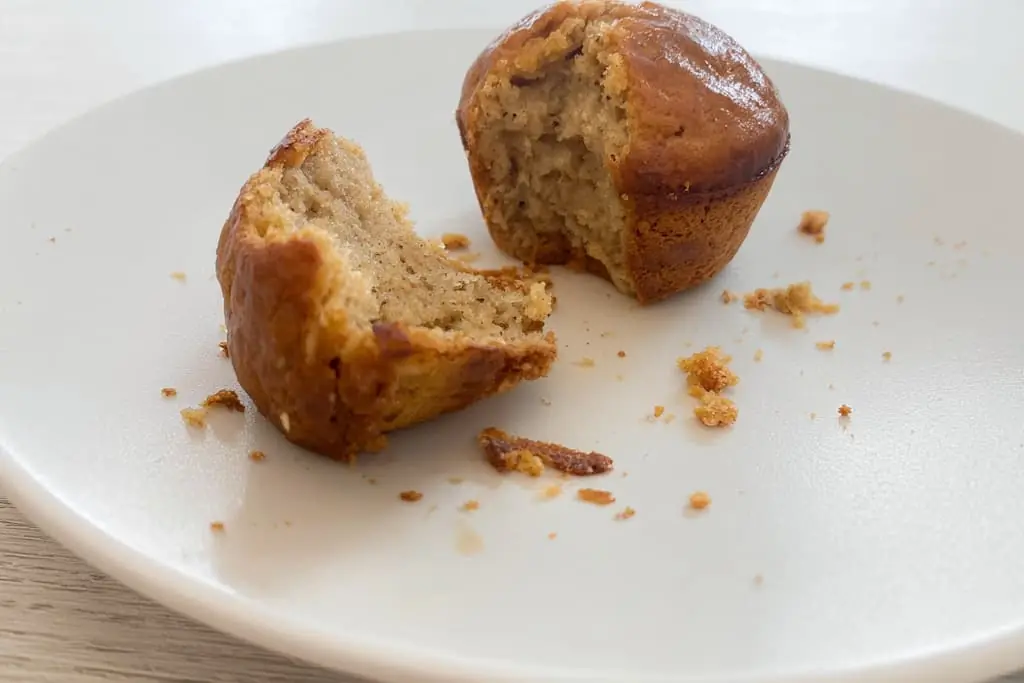 Cross-section view of a high protein banana muffin showing the tender, golden crumb structure with one half broken apart on a white plate with scattered crumbs