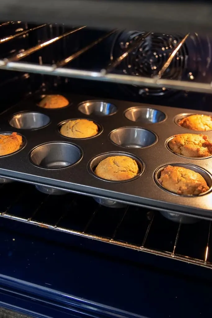 High protein banana muffins baking in a nonstick muffin tin inside an oven, showing various stages of doneness from risen golden brown to still baking