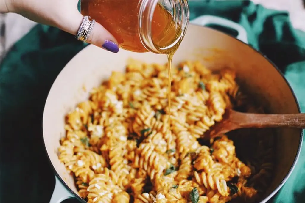Hand pouring golden honey from a small glass jar onto a pot of creamy rotini pasta with butternut squash, feta, and sage.