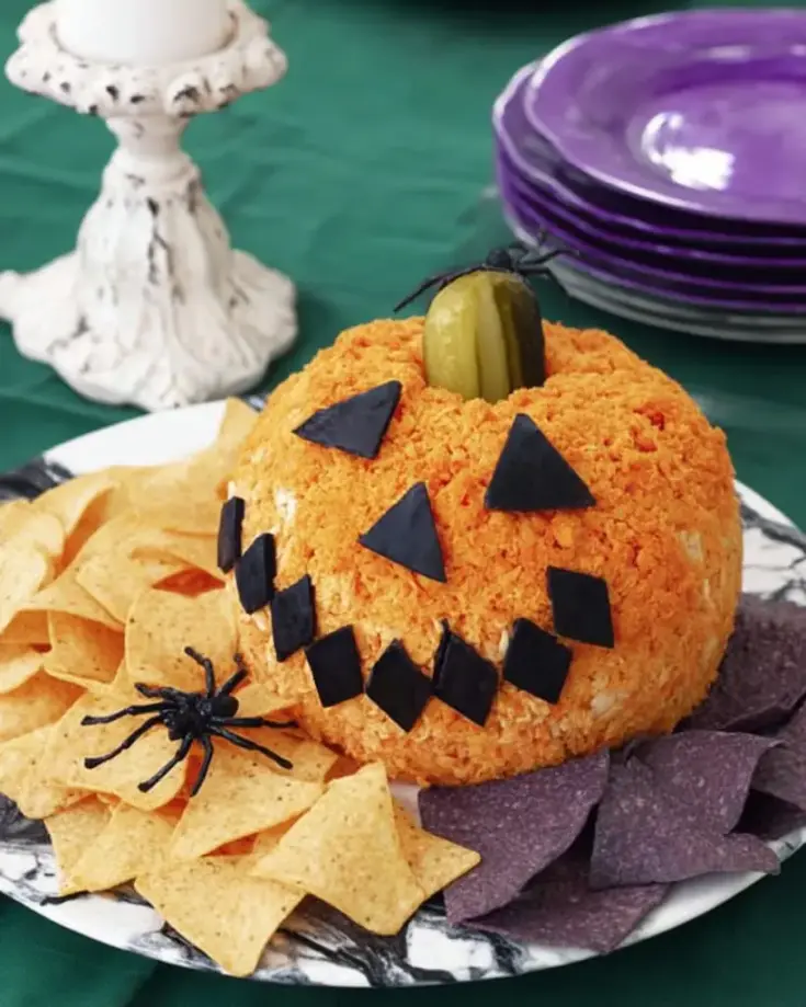Orange jack-o'-lantern cheese ball with black olive triangle eyes and zigzag mouth, topped with pickle stem, surrounded by tortilla chips on white serving plate