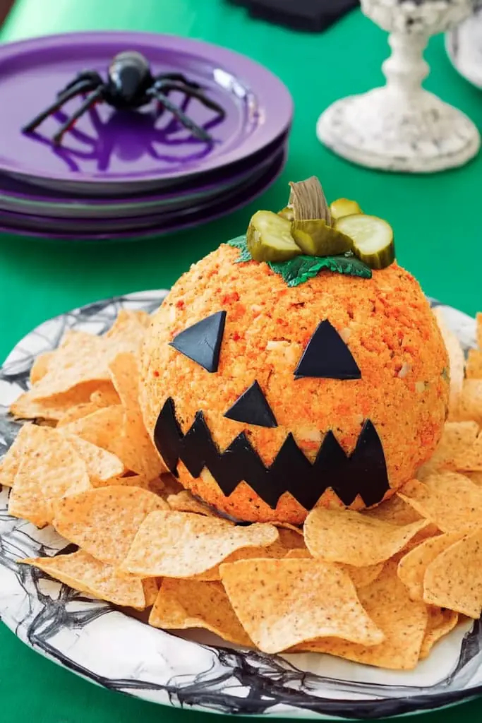 Orange jack-o'-lantern cheese ball with black olive triangle eyes and zigzag mouth, topped with sliced pickle stem, surrounded by tortilla chips on decorative plate with Halloween spider decorations
