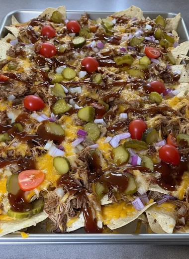 Sheet pan of crispy tortilla chips piled with smoky shredded pork, melted cheddar-jack cheese, dill pickle slices, halved cherry tomatoes, diced red onion, and a drizzle of thick barbecue sauce.